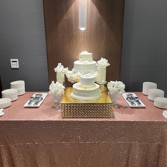 Wedding cake on a gold stand, flanked by floral arrangements and plates on a rose gold table.