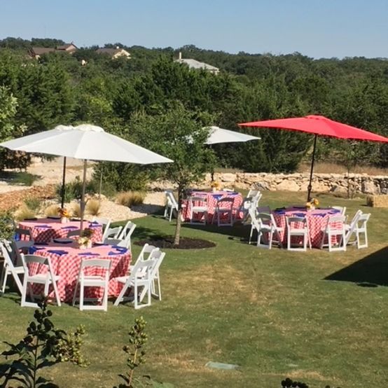 Outdoor tables with red and white checkered tablecloths and white umbrellas, on a grassy lawn.