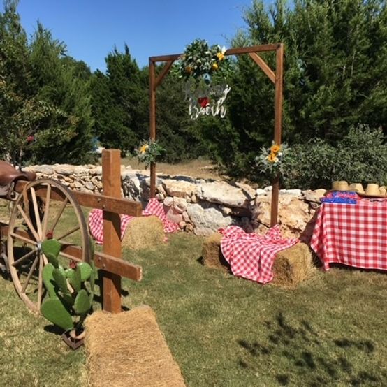 Rustic outdoor wedding setup with wooden arch, hay bales, and red checkered tablecloth.