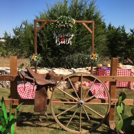 Western-themed photo booth with a wooden arch, saddle, wagon wheel, and picnic table.