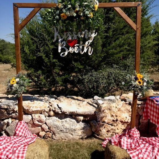 Wooden kissing booth with red gingham, floral accents, and hay bales outdoors.
