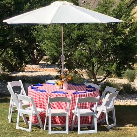 Round table with red checkered tablecloth, white chairs, umbrella in a garden.