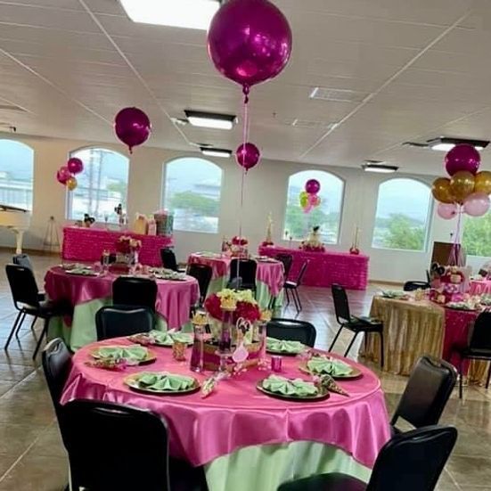 Tables set for a party with pink linens, balloons, and floral centerpieces. Indoors with windows.