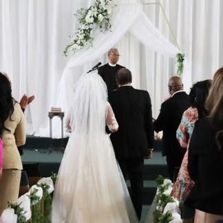 Bride walking down the aisle at a wedding ceremony, led by a man in a suit, with guests applauding.