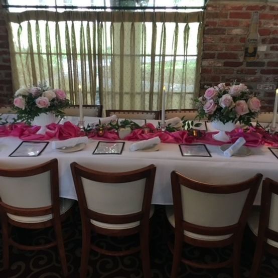 Wedding table set with pink roses, candles, and decorations; indoors with a window and brick wall.