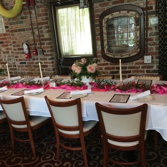 A formal dining table set for a celebration, decorated with pink ribbons, flowers, and candles, set in a brick-walled room.
