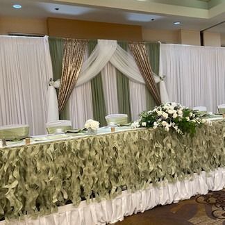 Head table at a wedding, sage green and white decor, draped fabric, floral arrangements.