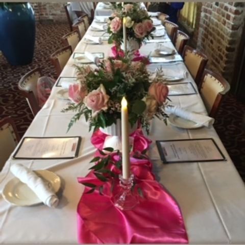 Long, white-tablecloth table set for a formal meal. Pink floral centerpieces, napkins, and menus.