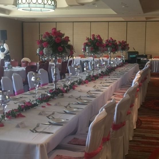 Long banquet table set for a formal event; white tablecloth, pink accents, red floral centerpieces, and white chair covers.