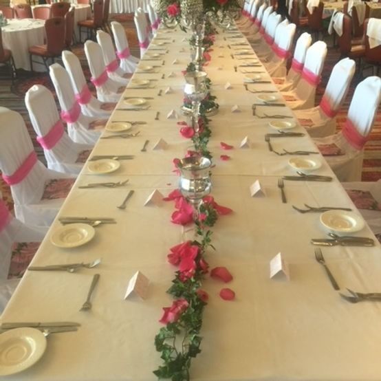 Long banquet table set with white linens, pink floral accents, and place settings.