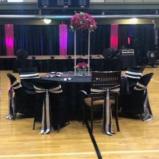 A decorated round table set for an event. Black tablecloth, chairs, with a floral centerpiece in a gymnasium.