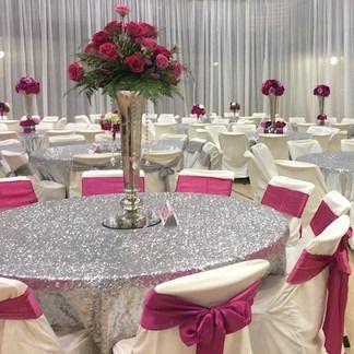 Elegant event hall with silver sequin tablecloths, pink floral centerpieces, and white chairs with pink bows.