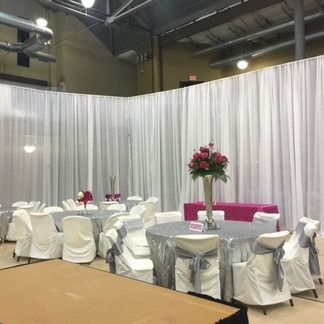 Event room with white draped walls, tables with silver linens, chairs, and floral centerpieces.