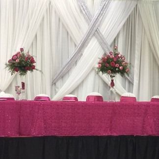 A decorated banquet table with magenta tablecloth, flowers, and white draped backdrop.