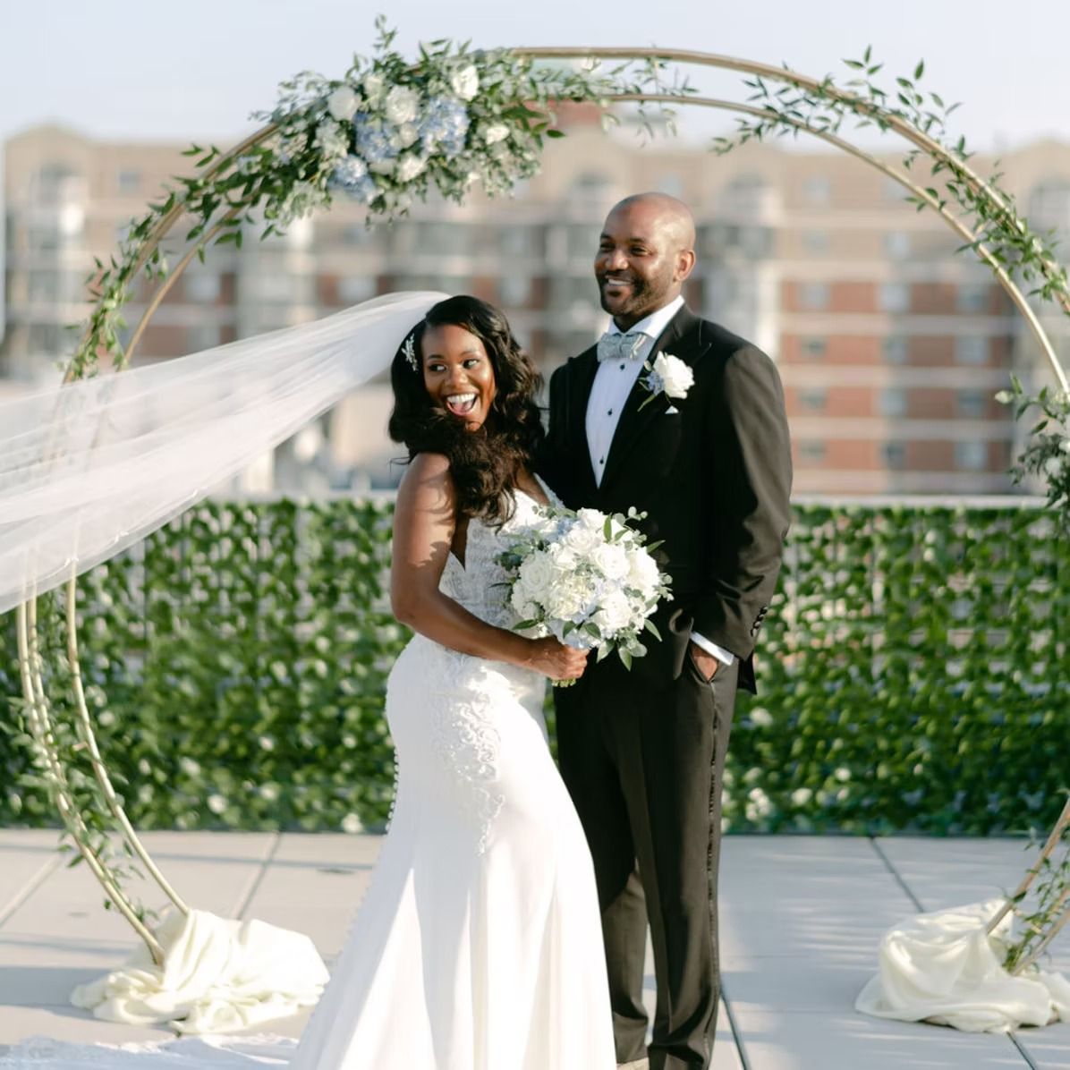 Newlyweds pose under a floral arch on a rooftop; bride wears a white gown, groom in a tuxedo.