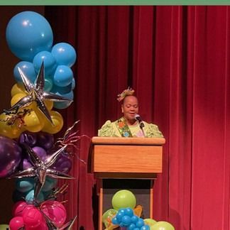 Woman speaking at podium with colorful balloons in front of red curtains.