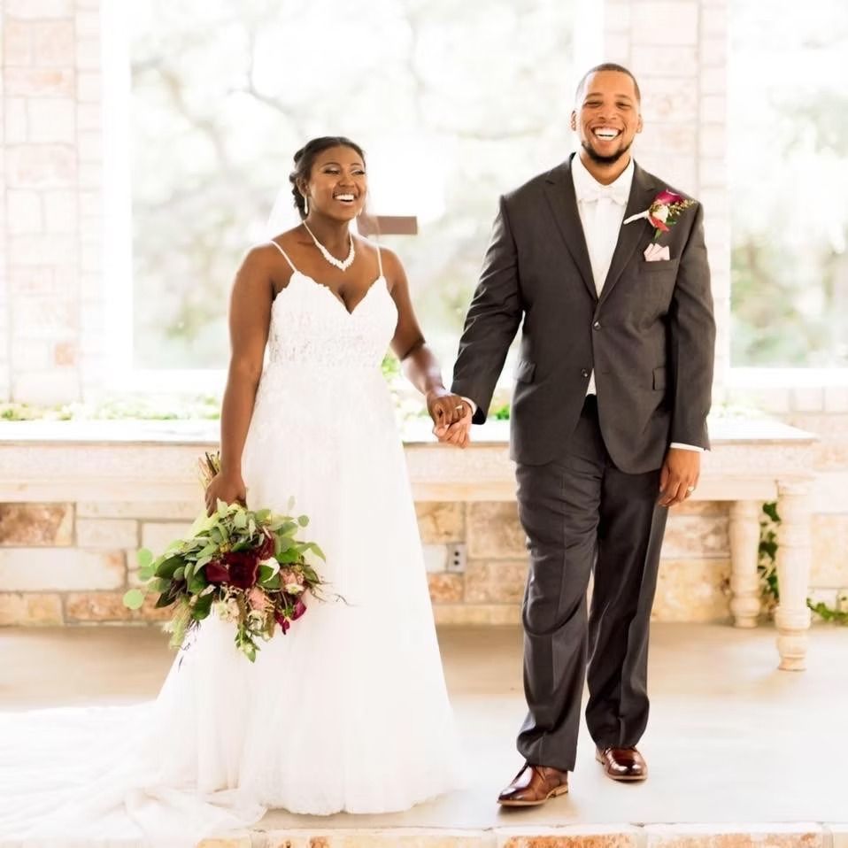 Newlyweds walk hand-in-hand smiling. The bride in white gown and bouquet; the groom in a gray suit. In a stone building.