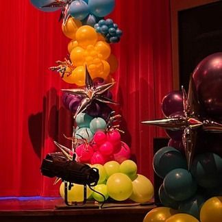 Colorful balloon decorations with starburst accents, a spotlight, and a red curtain.