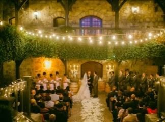 Wedding ceremony in stone courtyard, bride and groom at altar, guests seated, fairy lights.