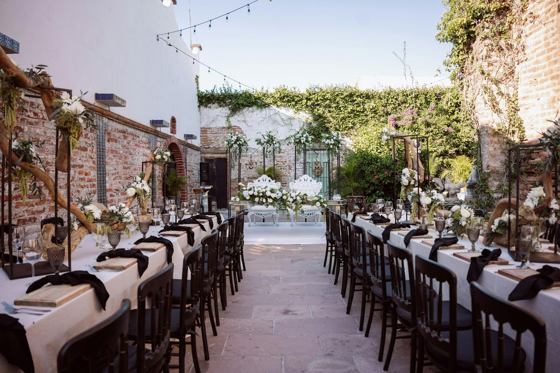 Long dining tables set for a wedding in a courtyard, with black chairs and floral arrangements.