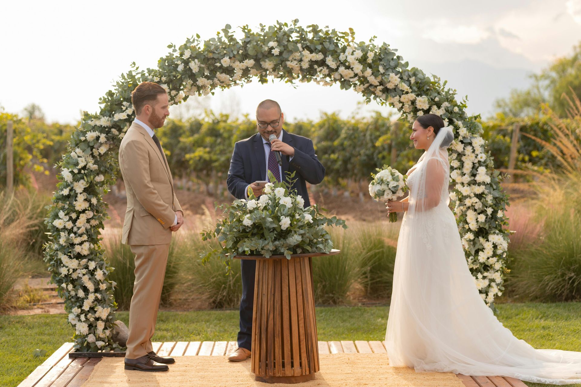 Wedding ceremony with bride, groom, officiant under a floral arch. Vineyard backdrop, sunny day.