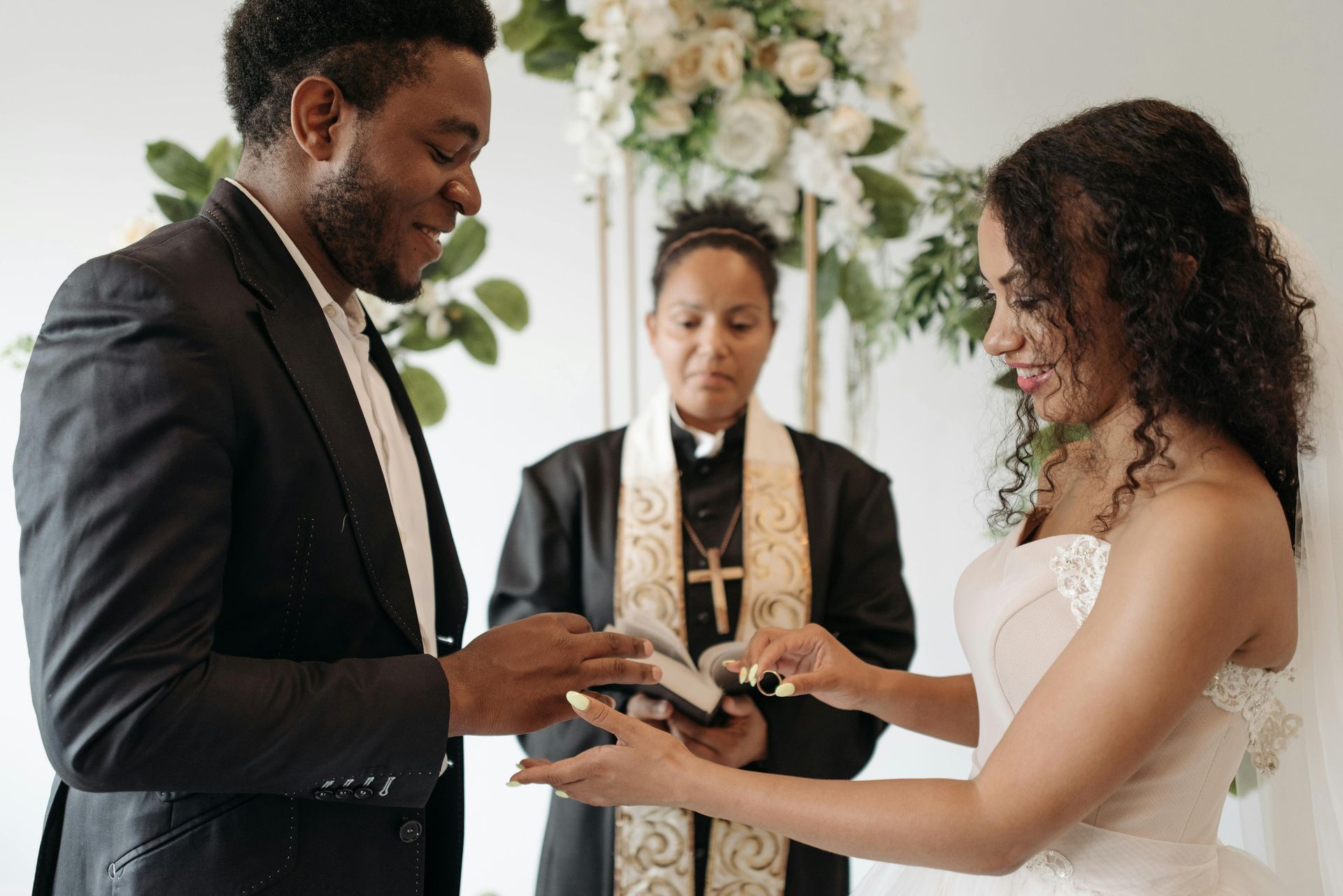 Groom placing ring on bride's finger during a wedding ceremony, officiant in background.