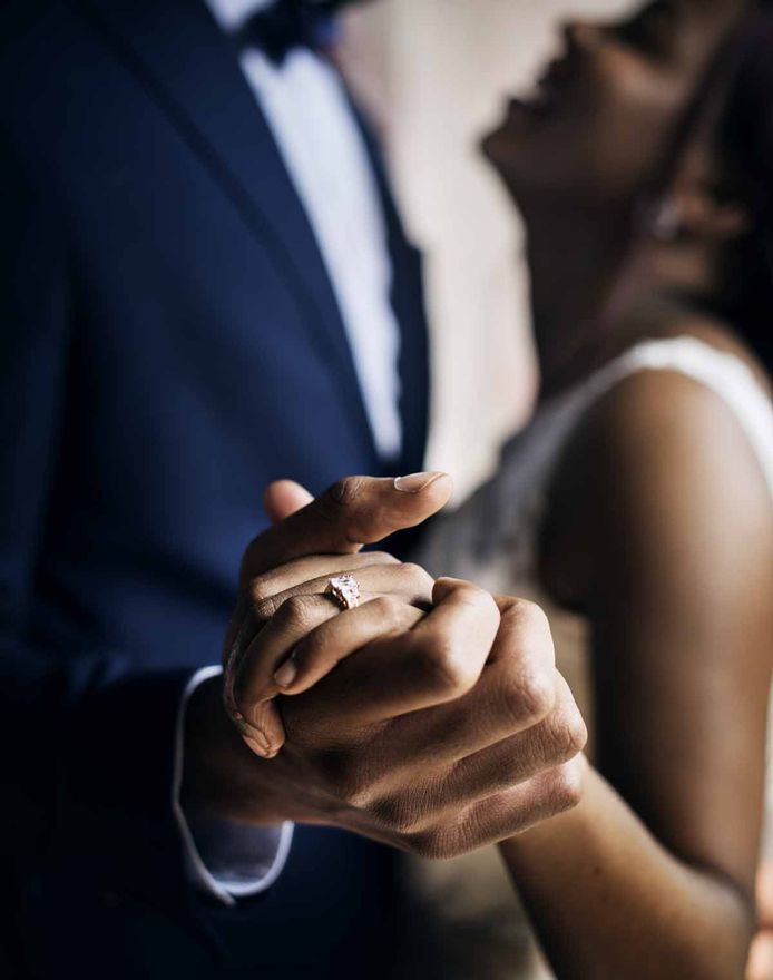 Couple dancing, close-up of hands clasped; woman's ring visible. Man in navy suit. Silhouette kiss in the background.
