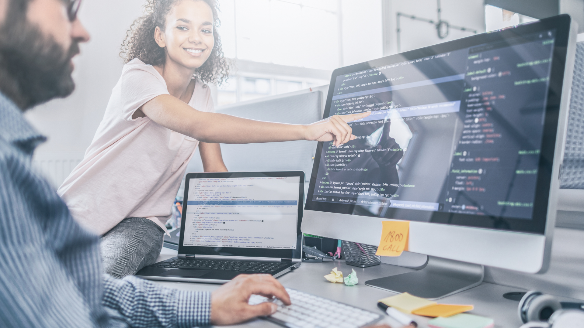 Woman pointing at computer screen with code, next to a man at a desk, laptop visible.