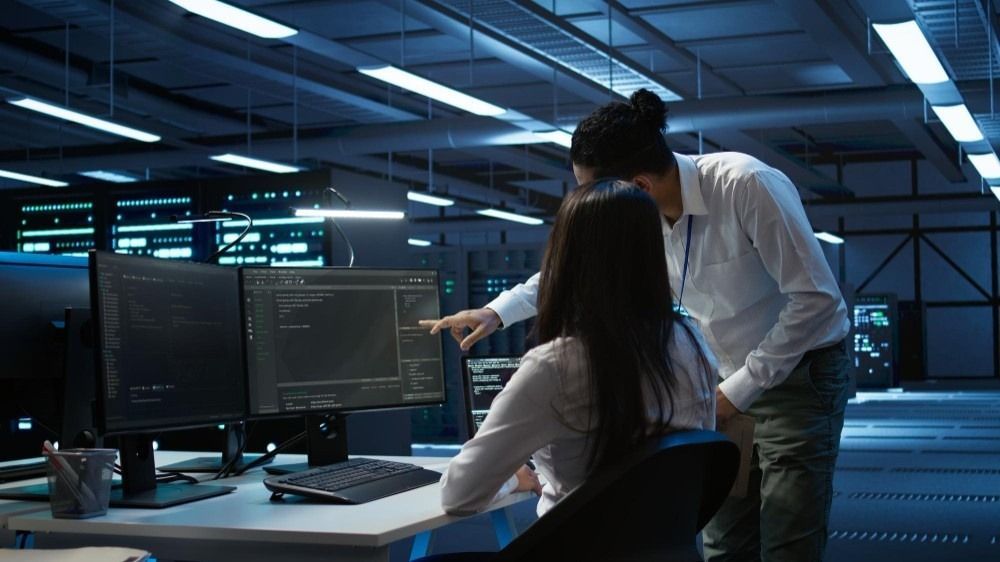 Two people in data center, pointing at computer screens, discussing code. Blue lighting.