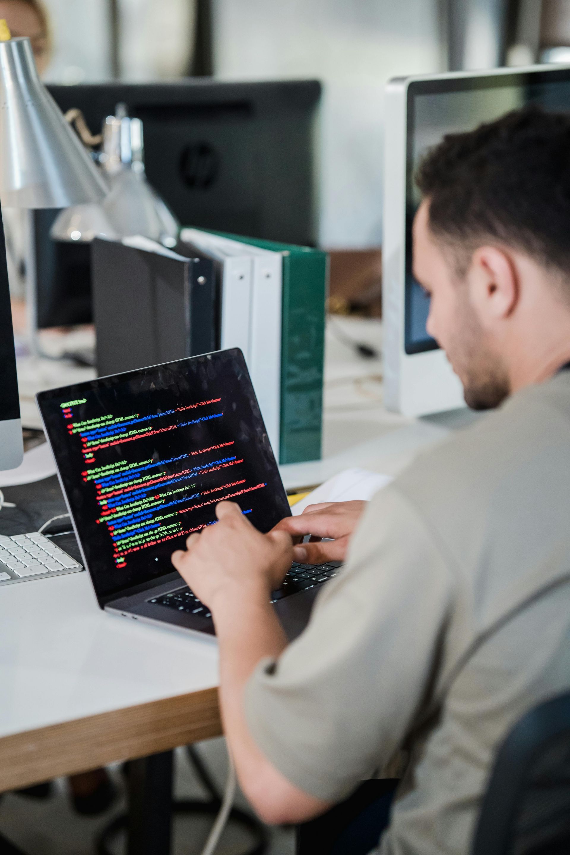 Man working on a laptop, coding in a bright office. The screen displays colorful code; he's pointing at it.