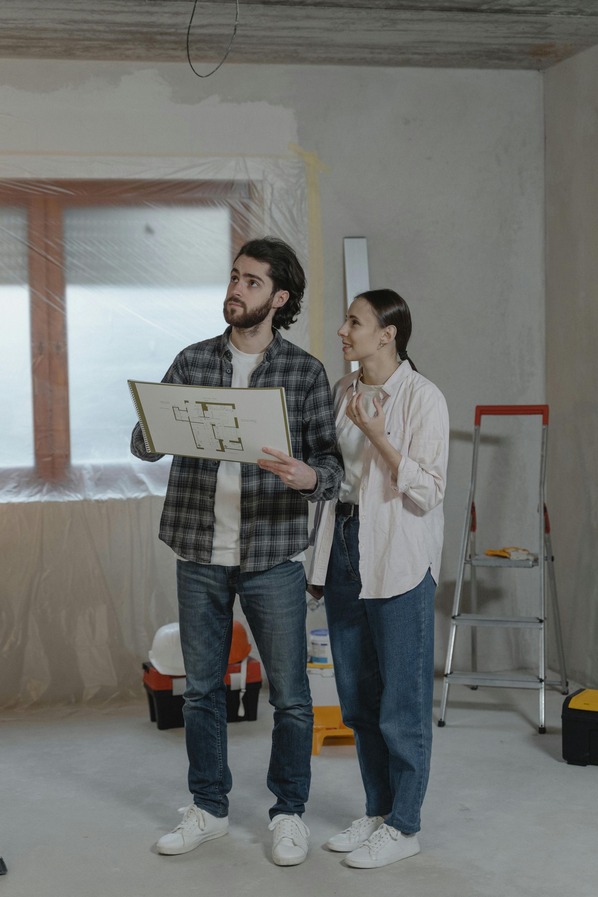 A couple reviewing a blueprint in a room under renovation. The man holds the plan, while the woman gestures, both looking upwards.