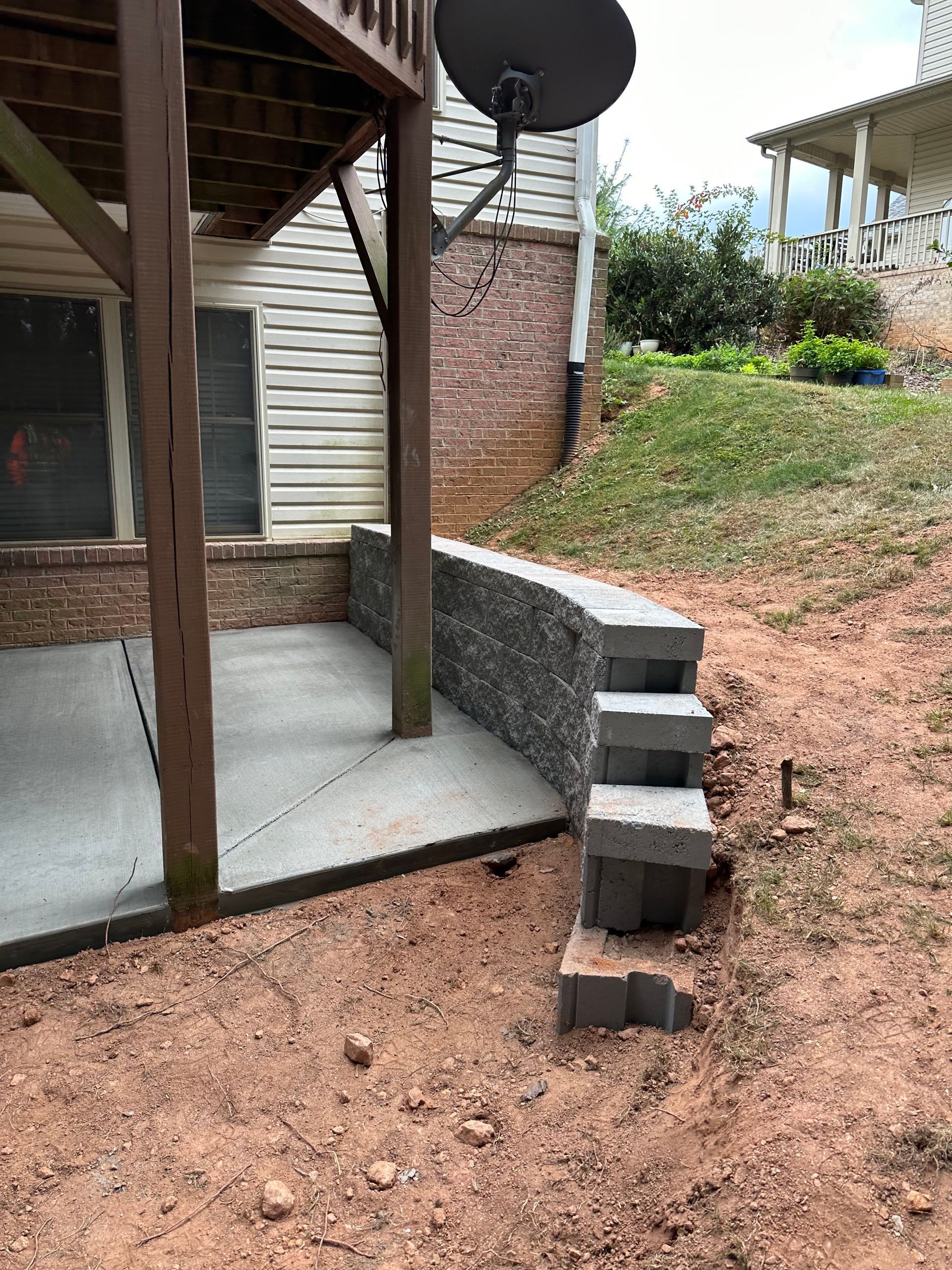 Concrete patio with a curved stone retaining wall and steps leading to a sloped grassy backyard.