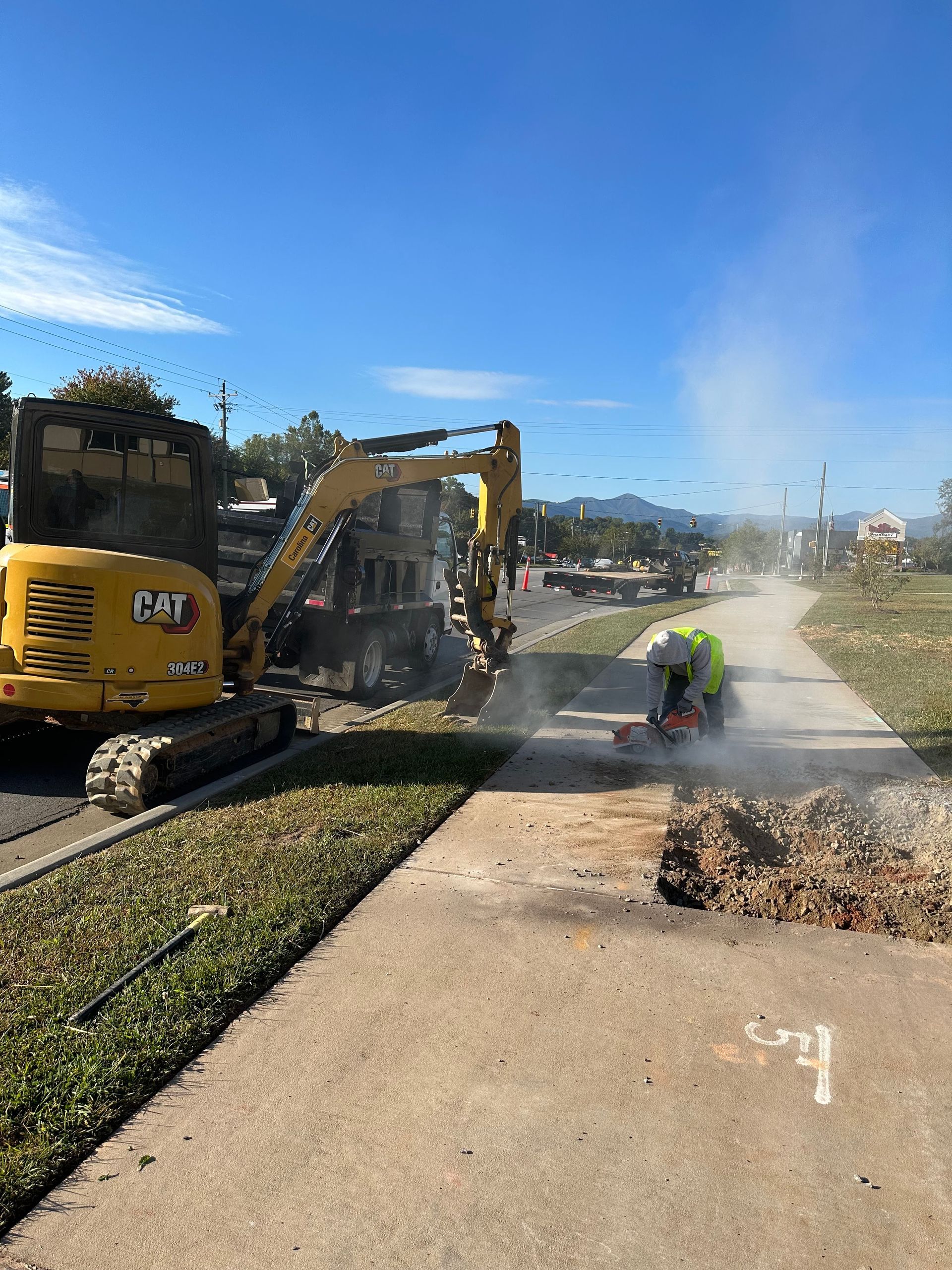 A construction worker uses a power saw on a sidewalk near a yellow CAT excavator under a bright blue sky.