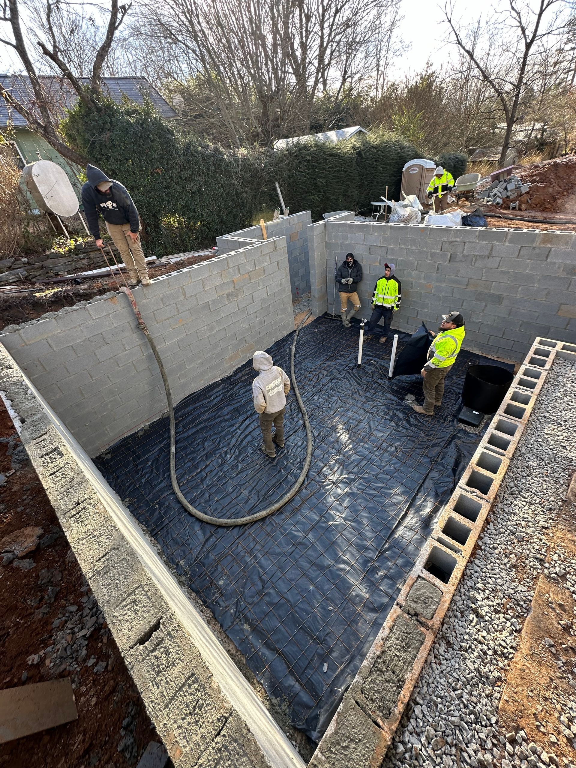 Construction workers standing in a concrete foundation pit with a black waterproof liner on the ground.