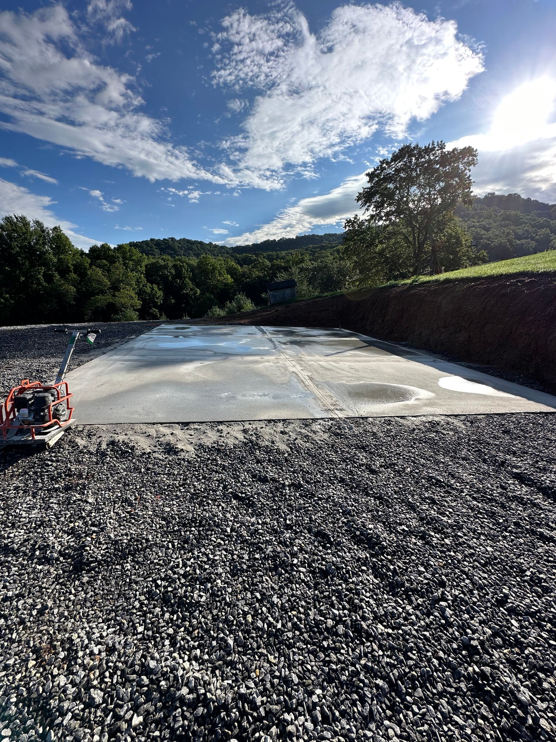 A newly poured rectangular concrete pad sits on a gravel lot under a bright blue sky with hills in the background.