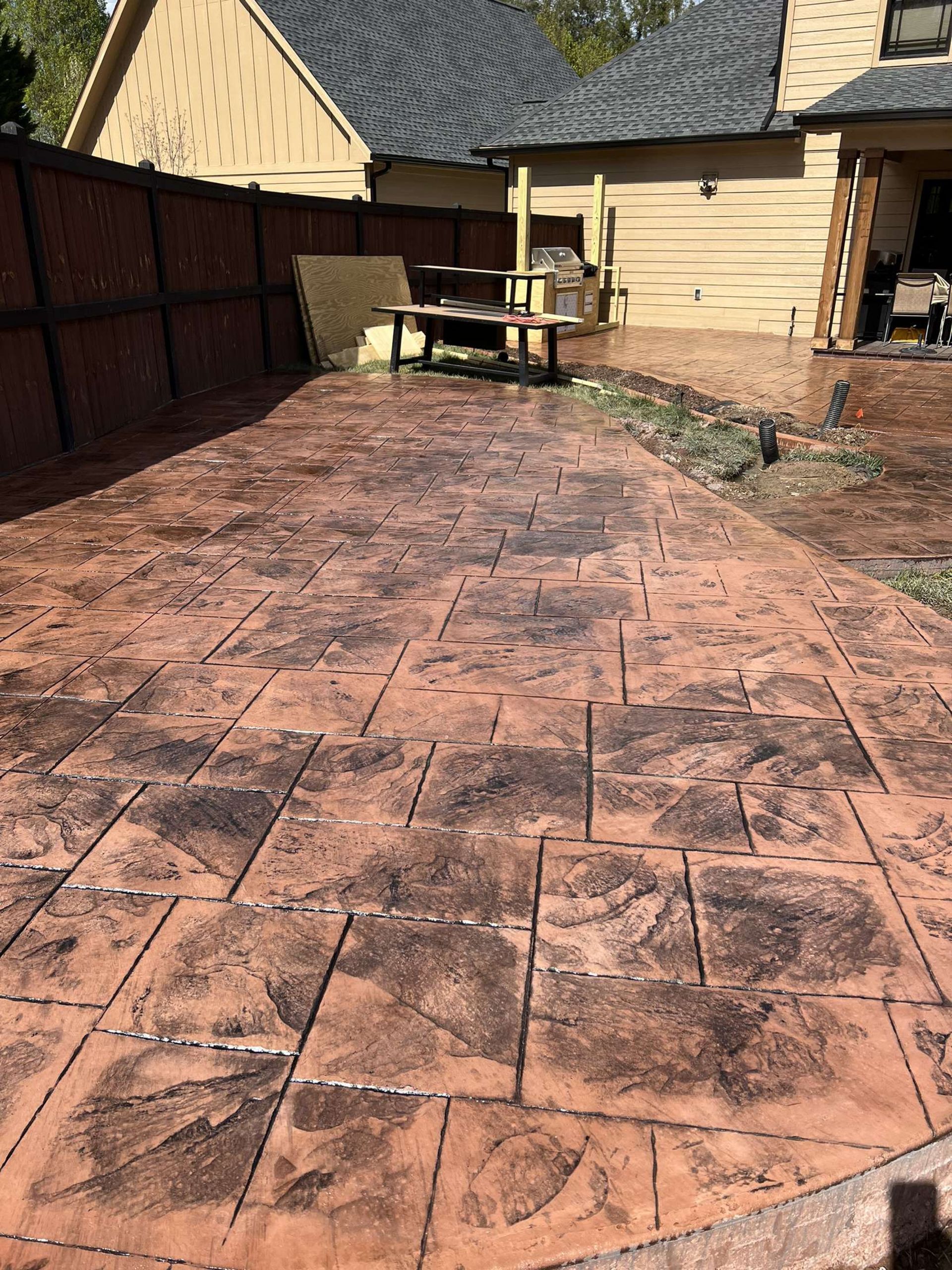 A backyard patio with stamped, reddish-brown concrete tiles, a dark wooden fence, and a beige house in the background.