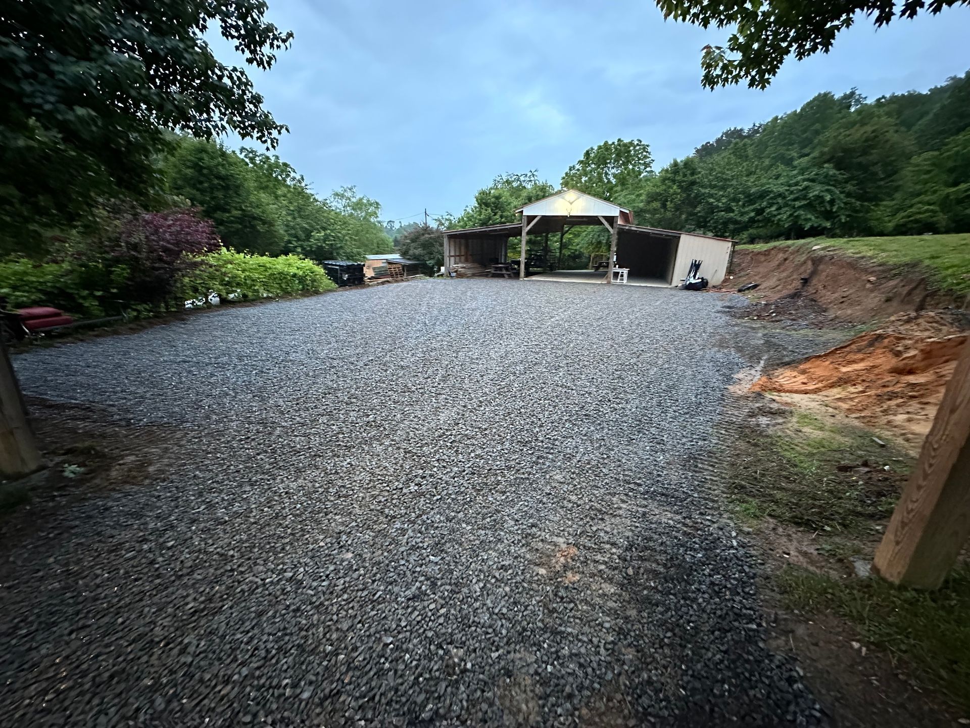 A wide gravel driveway leads to a wooden open-sided barn surrounded by trees under a cloudy sky.