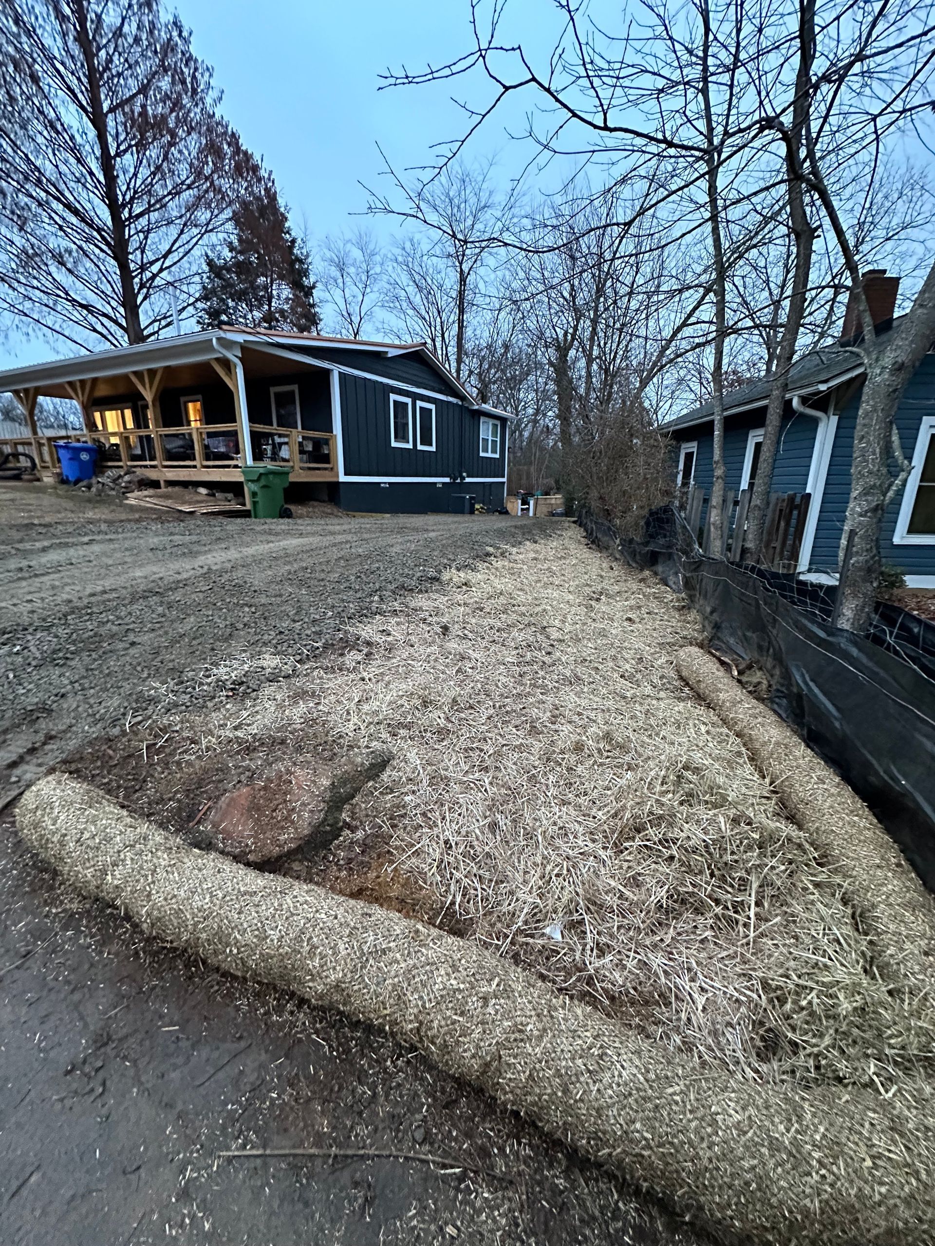 A dark blue house with a wooden porch overlooks a gravel-covered yard with a decorative log-lined planting bed.