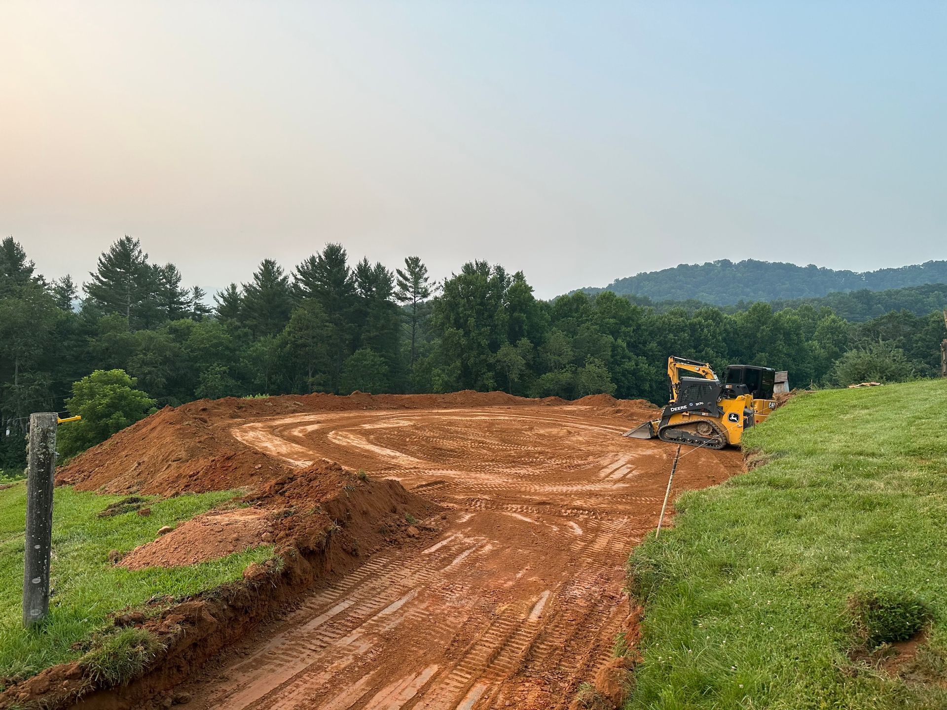 A yellow bulldozer levels red clay dirt on a construction site next to a grassy hill and a line of green trees.