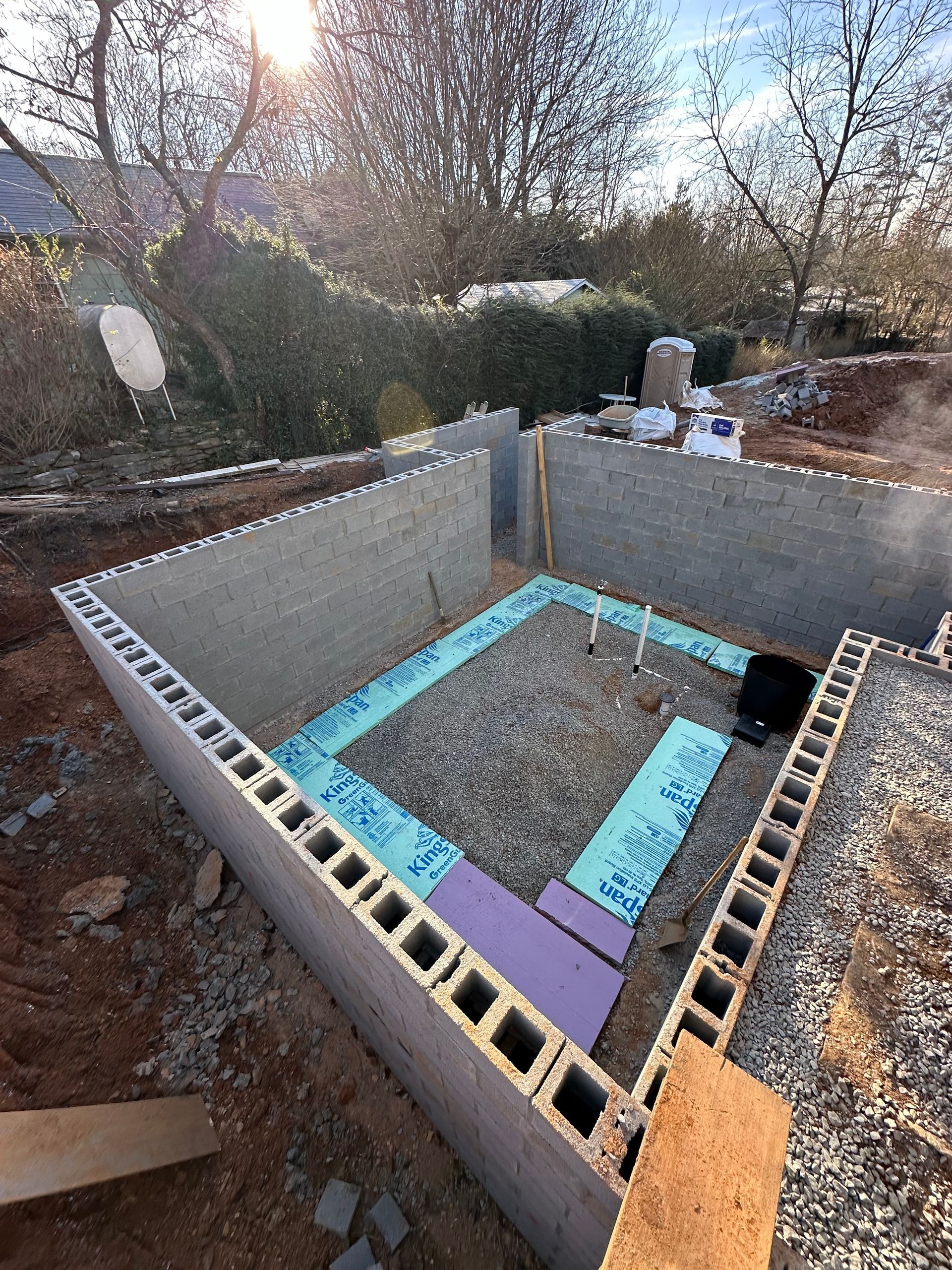 A construction site showing concrete block foundation walls and insulation boards on a gravel base under a bright sun.