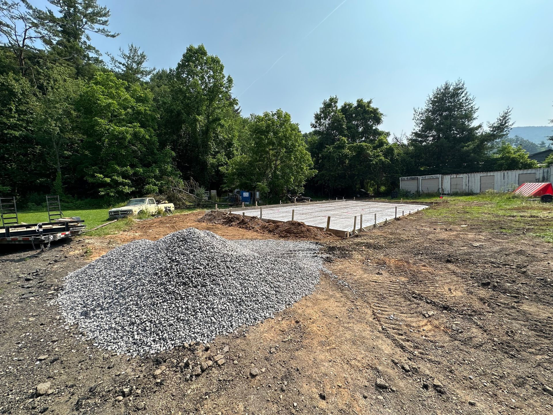 A construction site showing a concrete foundation slab, a large pile of gravel, and surrounding trees under a blue sky.