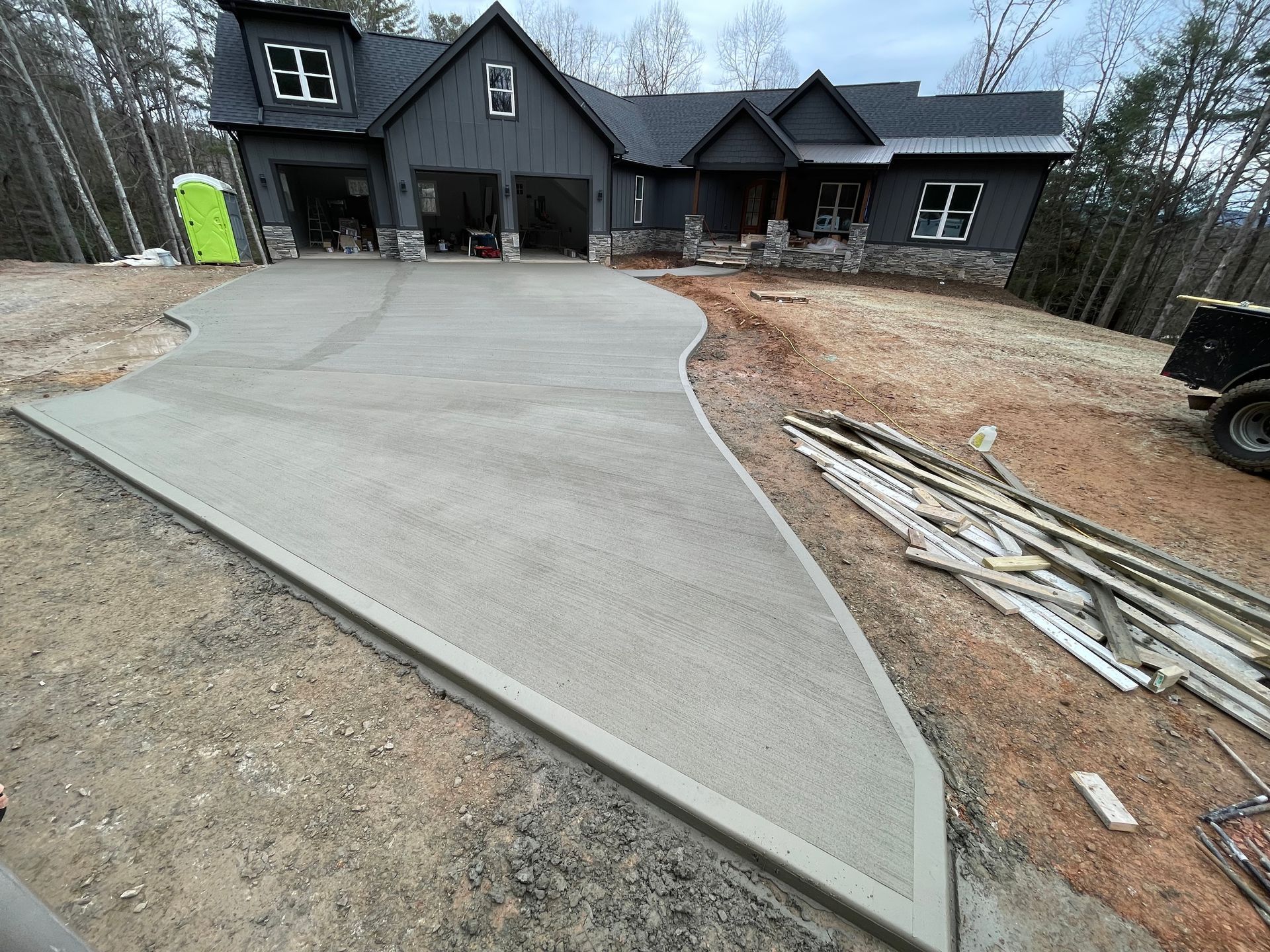 A new concrete driveway leads to a dark, modern house under construction in a wooded area.