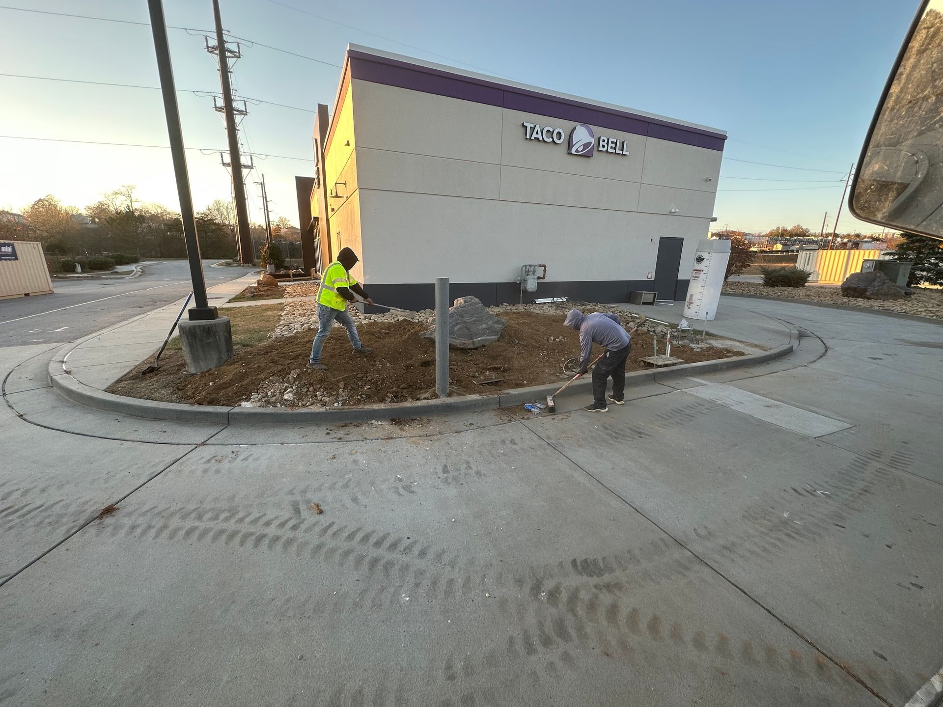 Two workers in bright safety vests landscape a dirt patch in front of a Taco Bell building on a sunny day.