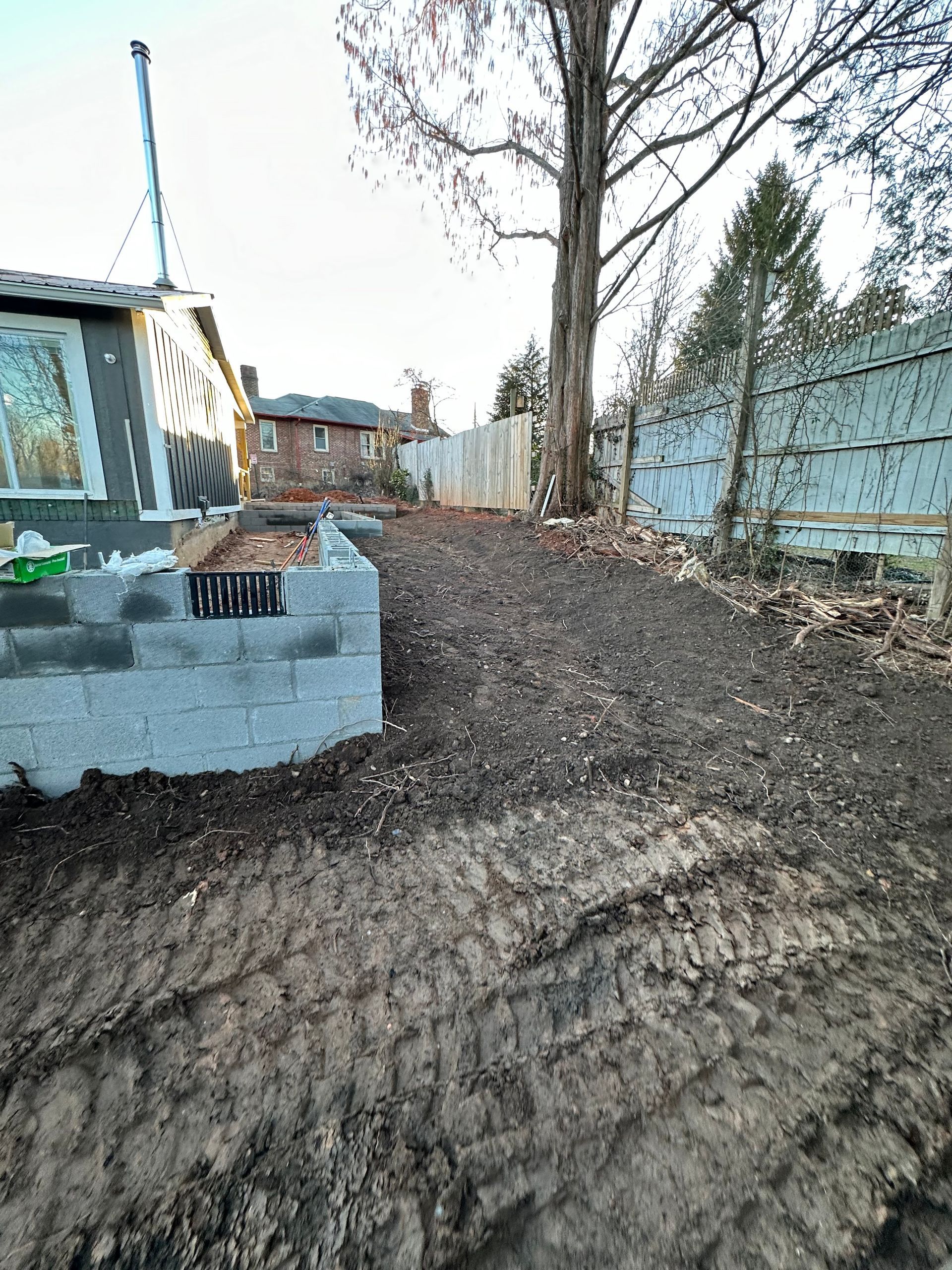 A concrete block wall under construction in a yard with fresh soil, tire tracks, and a house in the background.