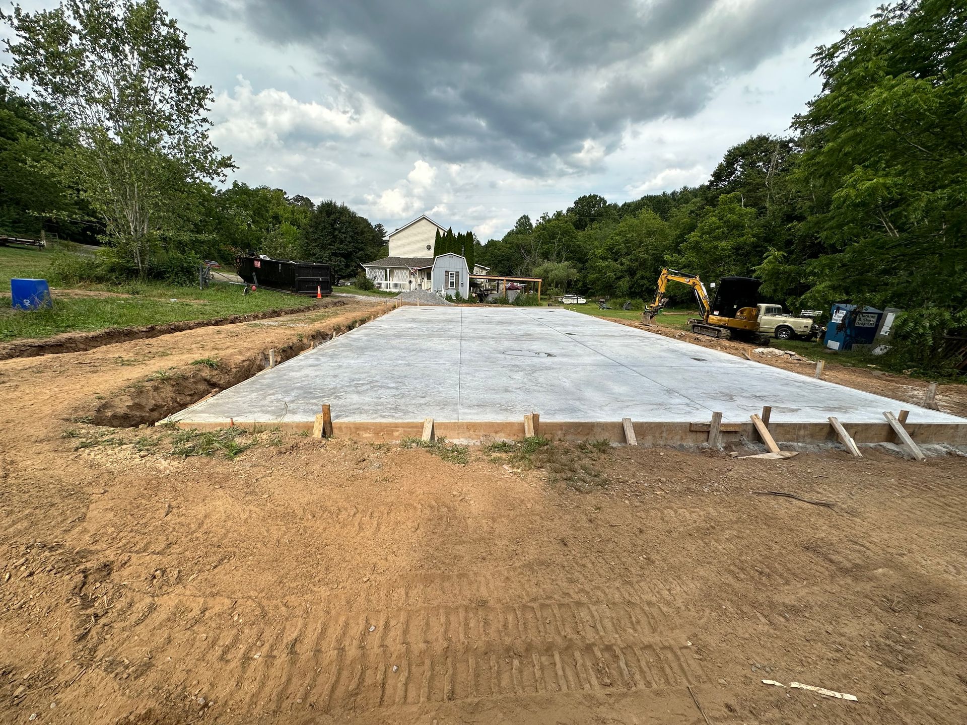 A newly poured, rectangular concrete slab foundation in a grassy, wooded construction site under a cloudy sky.