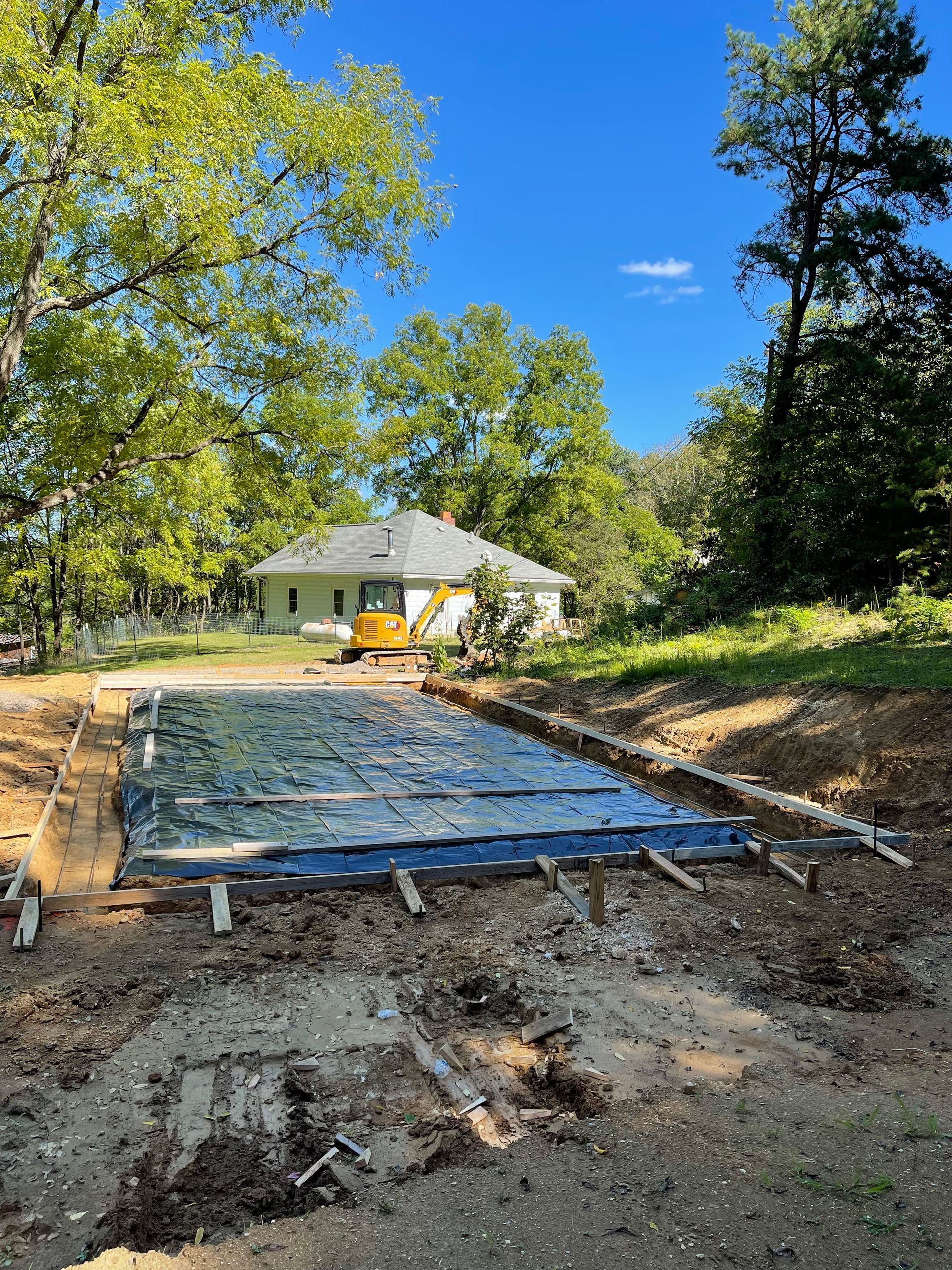 Construction site showing a rectangular foundation covered with a black tarp, framed by wooden forms on a sunny day.