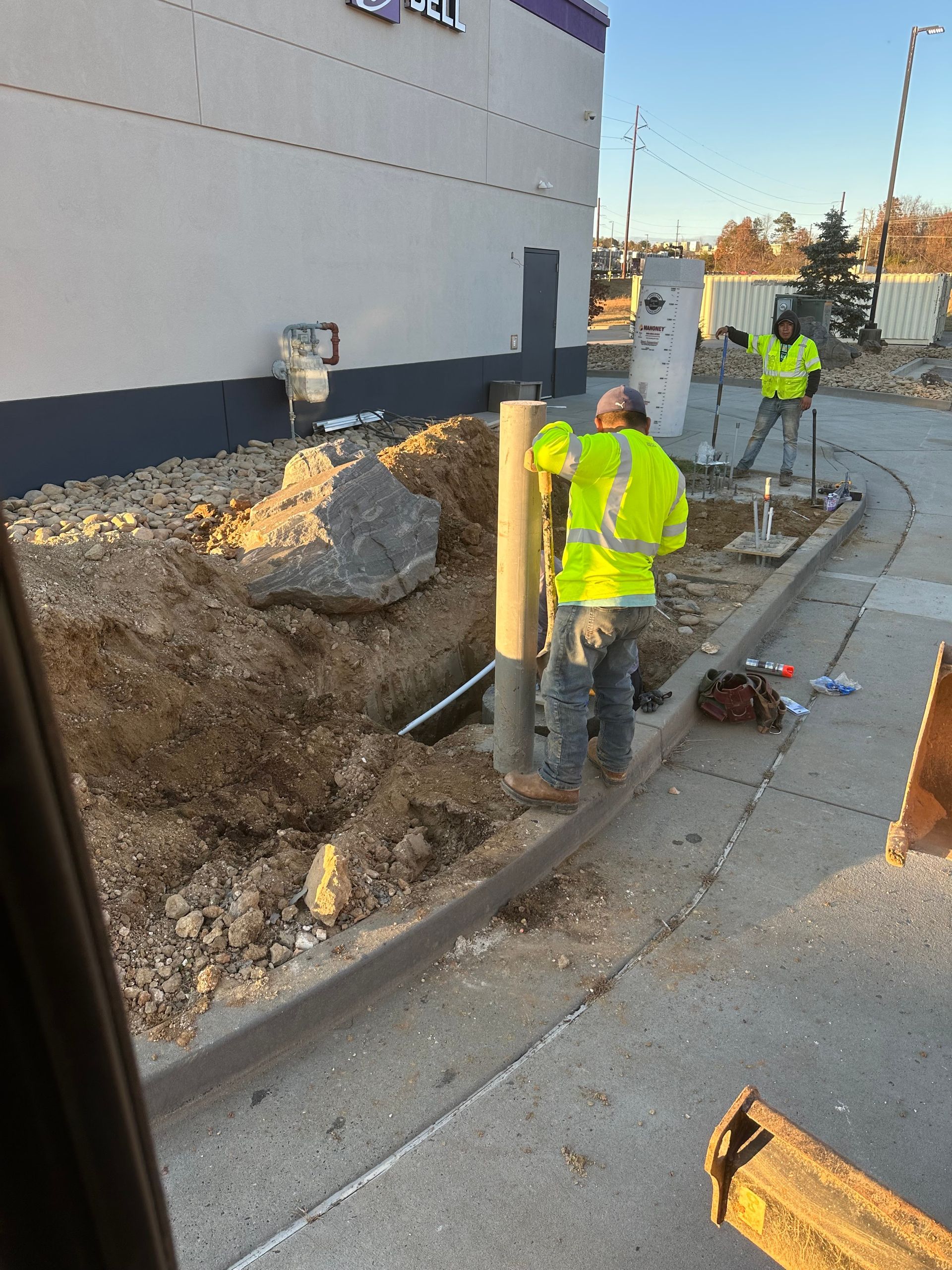 Two construction workers in neon yellow safety gear working on a trench excavation beside a building.