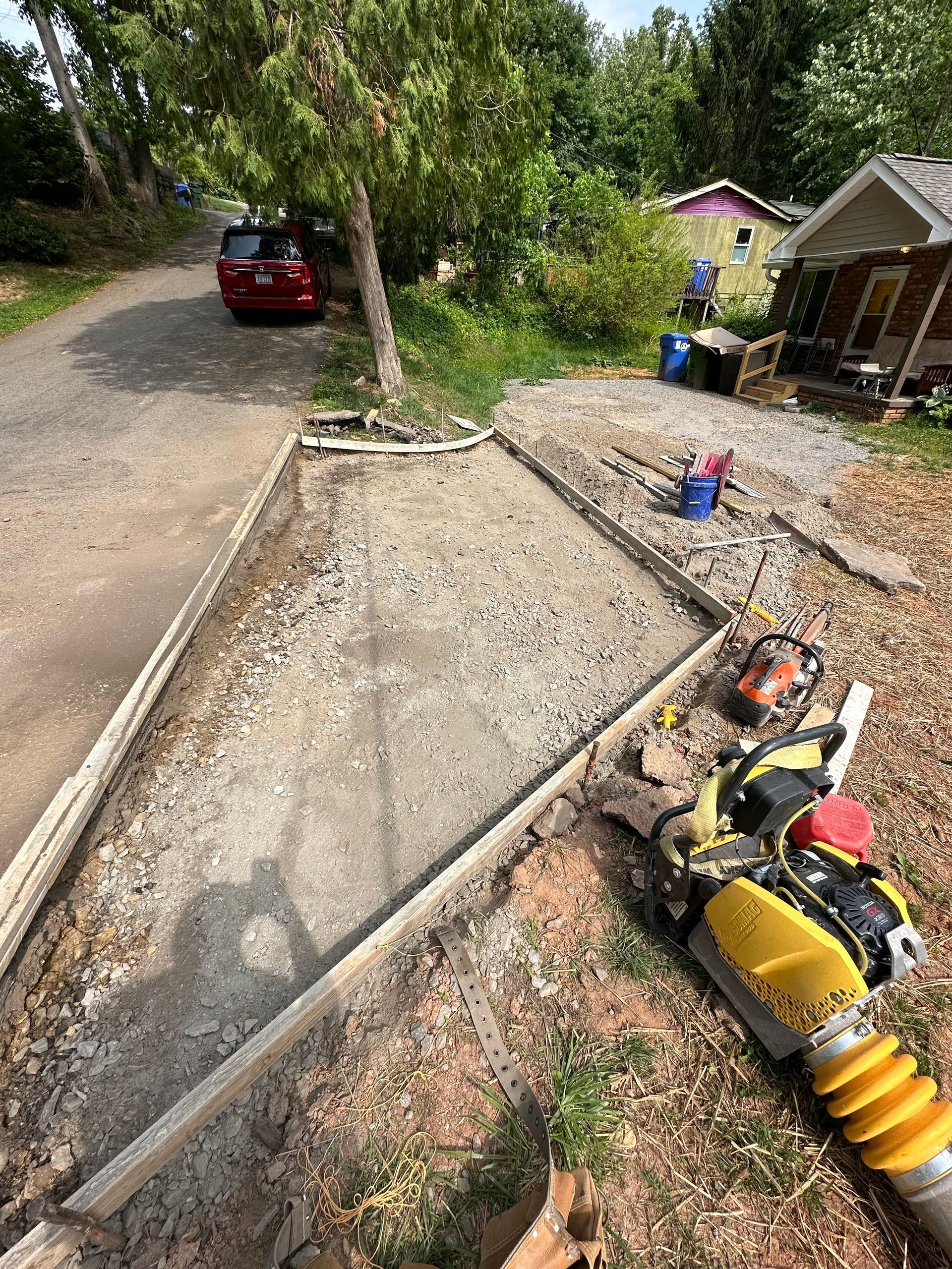 A gravel driveway base under construction, framed by wooden forms, with a yellow plate compactor sitting nearby.