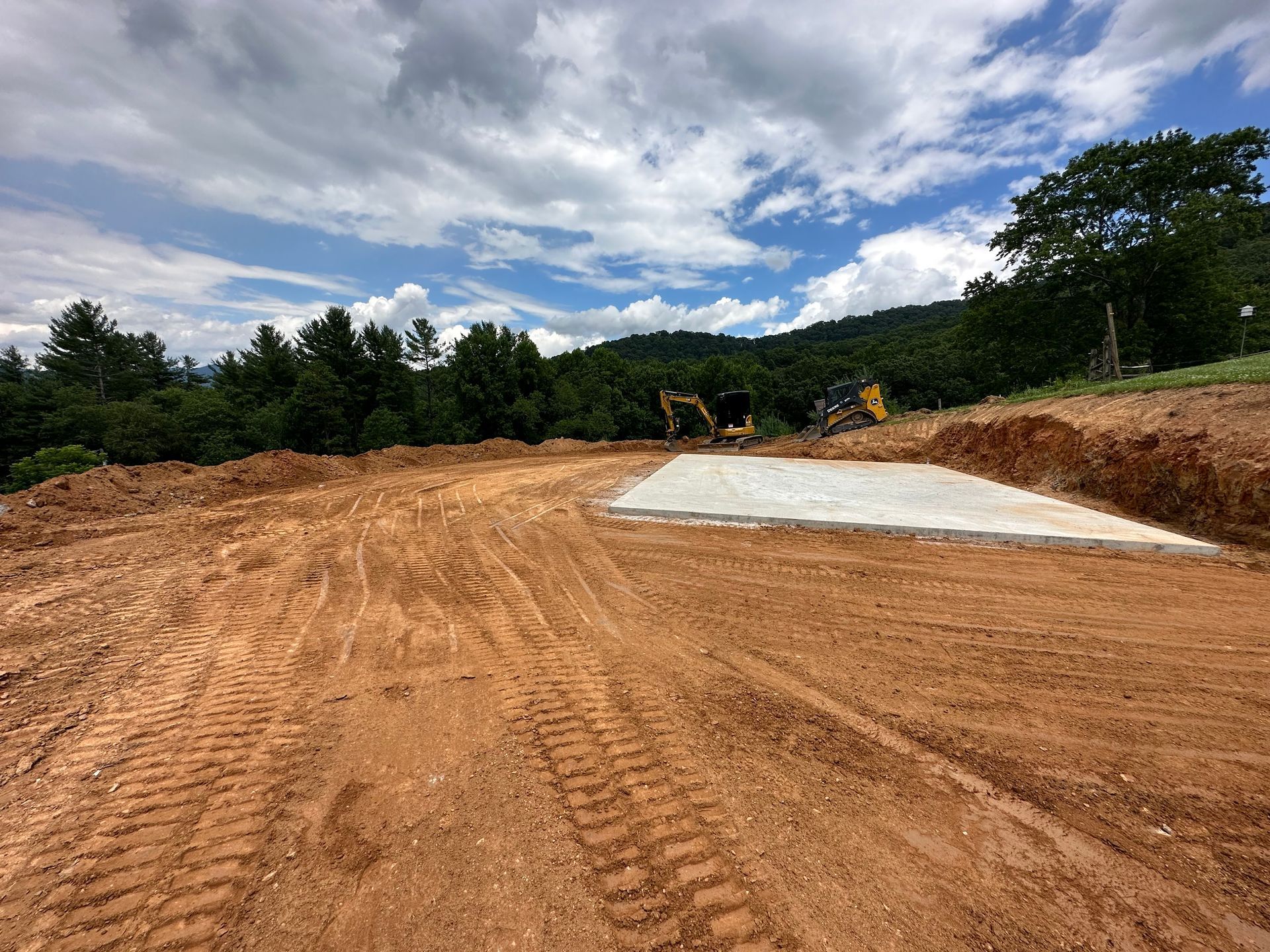 A concrete foundation pad sits on graded earth at a construction site with trees and mountains in the background.