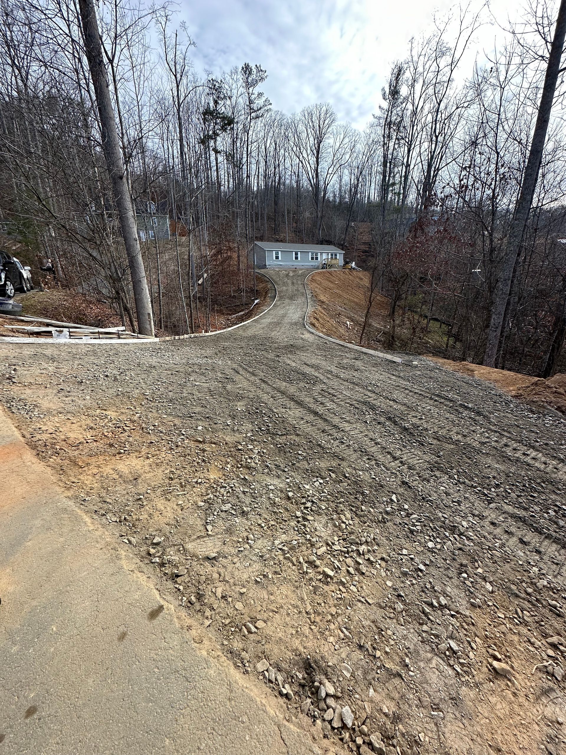 A gravel driveway leading from a paved road up a gentle slope toward a house nestled among bare trees in the distance.
